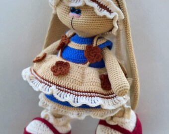 May include: A crocheted rabbit doll with long ears, wearing a blue and beige dress adorned with brown flower appliqués and white ruffled trim. The doll features blue bead eyes, a pink stitched nose, and wears cream-colored shoes with red soles.