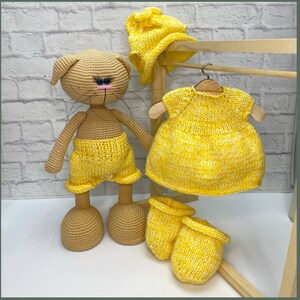 Op de afbeelding: Een gehaakte kattenpop die een gele gebreide outfit draagt, inclusief een broekje, een jurk, een muts en slofjes. De pop staat op een houten oppervlak.