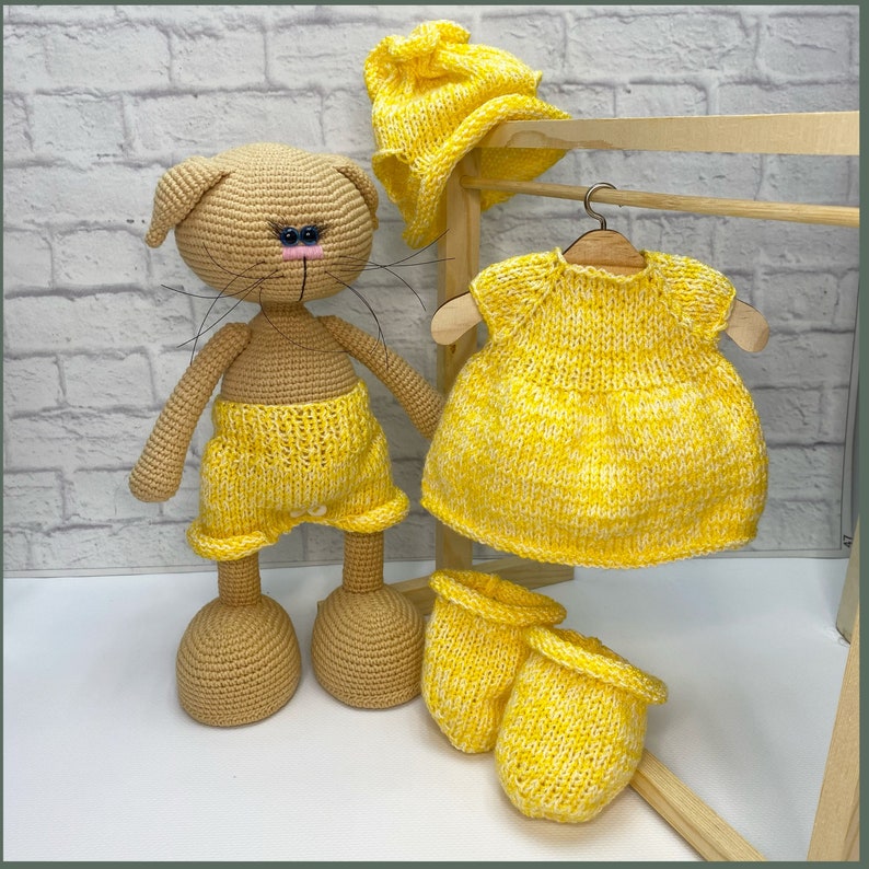 Op de afbeelding: Een gehaakte kattenpop die een gele gebreide outfit draagt, inclusief een broekje, een jurk, een muts en slofjes. De pop staat op een houten oppervlak.
