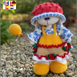 Könnte beinhalten: Eine gehäkelte Hasenpuppe, die einen rot-weißen Hut, ein blau-weiß gestreiftes Hemd, ein gelbes Latzkleid mit rot-weißem Besatz und gelbe Schuhe trägt. Der Hase hält einen gelben Ballon.