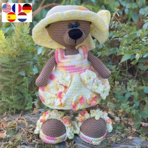 May include: A crocheted teddy bear wearing a yellow and pink dress with white flowers, a yellow sun hat, and brown sandals. The bear is standing on a bed of green leaves.