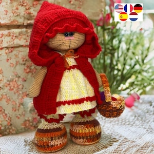 May include: A crocheted cat doll wearing a red hooded cape and a yellow and white dress. The doll is holding a brown basket.