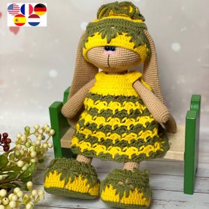 Könnte beinhalten: Eine handgefertigte Häkelkaninchenpuppe mit einem gelb-grünen Kleid, passendem Hut und Schuhen. Die Puppe hat lange Ohren und sitzt auf einer kleinen grünen Bank. Das Gesicht des Kaninchens ist beige mit blauen Augen und einer rosa Nase.
