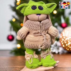Könnte beinhalten: Ein gehäkeltes Plüschtier, das als Figur aus dem Star Wars-Franchise verkleidet ist. Das Spielzeug trägt einen grünen Hut mit großen Ohren, einen braunen gehäkelten Mantel und grüne Schuhe. Das Spielzeug steht auf einer Hand.