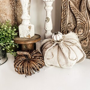 May include: A white fabric pumpkin with a white knob on top, a brown woven pumpkin, and a white wooden finial on a wooden base.
