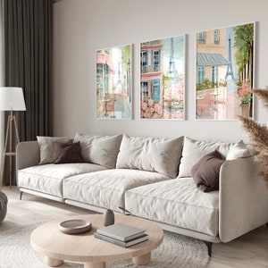 May include: Three framed watercolor paintings depicting Parisian street scenes with cafes, flowers, and the Eiffel Tower. The paintings are hung on a white wall in a living room setting with a white sofa, a coffee table, and a lamp.