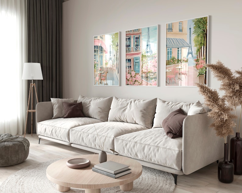 May include: Three framed watercolor paintings depicting Parisian street scenes with cafes, flowers, and the Eiffel Tower. The paintings are hung on a white wall in a living room setting with a white sofa, a coffee table, and a lamp.