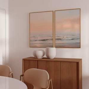 May include: Two framed beach scene prints with gold frames hang above a wooden cabinet. Two white, rounded vases sit on the cabinet. The prints depict a sunset over the ocean with surfers in the water.