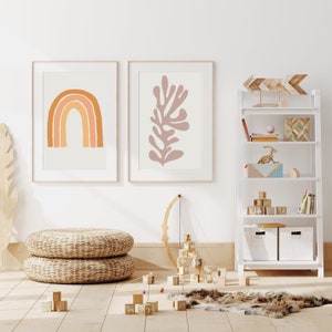 May include: Two framed art prints with abstract designs. One features a rainbow in shades of orange, the other a coral-like shape in mauve. A white bookshelf displays decorative items. Woven poufs and wooden blocks are on the floor.