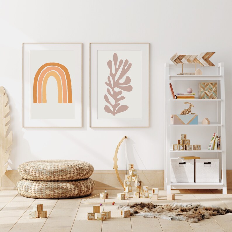 May include: Two framed art prints with abstract designs. One features a rainbow in shades of orange, the other a coral-like shape in mauve. A white bookshelf displays decorative items. Woven poufs and wooden blocks are on the floor.