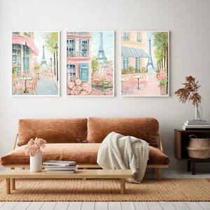 May include: Three watercolor paintings of Parisian street scenes featuring pink buildings, cafes, and the Eiffel Tower. The paintings are framed in white and hung on a white wall.