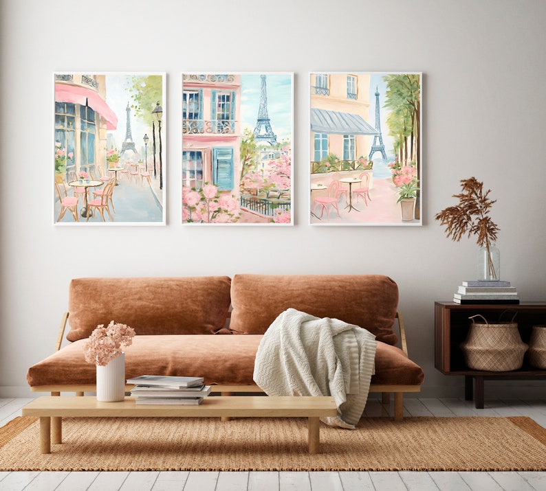 May include: Three watercolor paintings of Parisian street scenes featuring pink buildings, cafes, and the Eiffel Tower. The paintings are framed in white and hung on a white wall.
