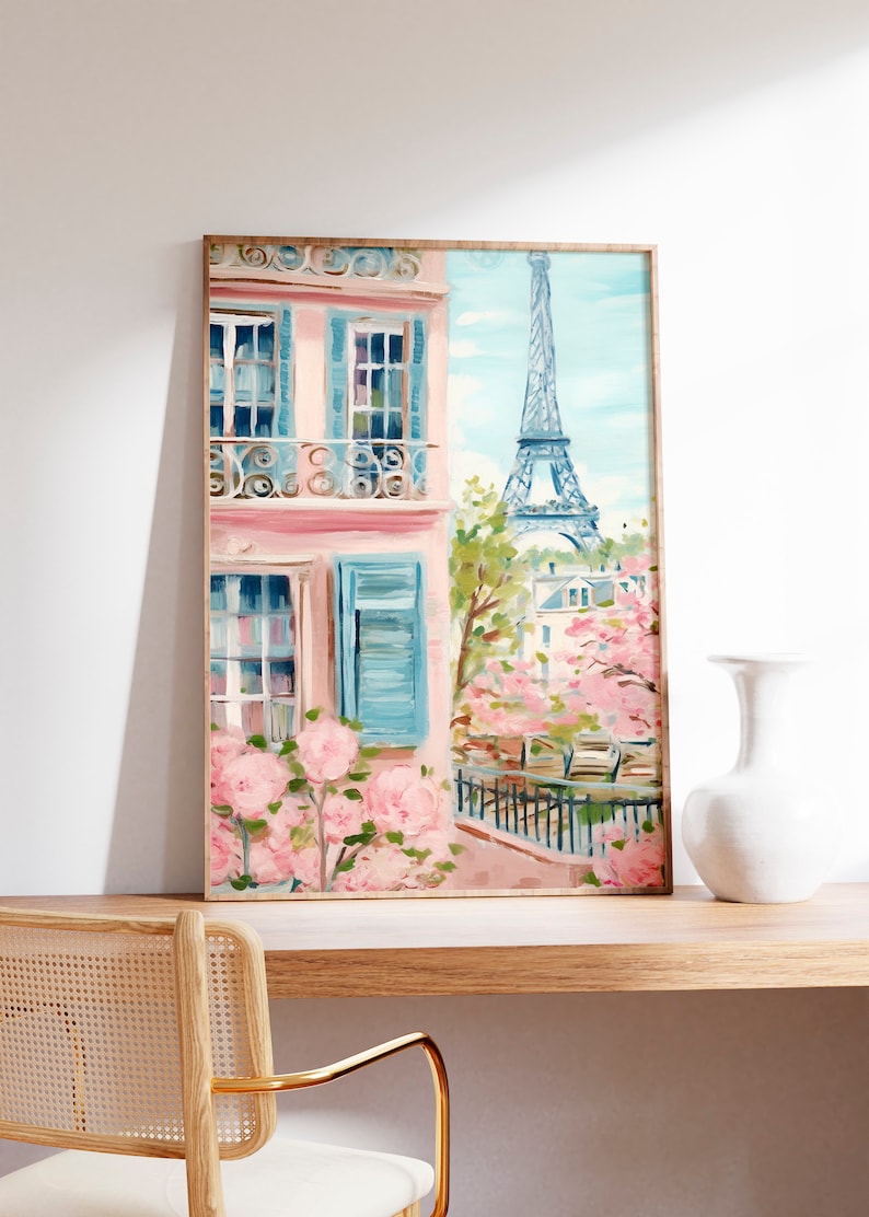Pu&ograve; includere: Un dipinto incorniciato di un edificio rosa con persiane blu e un balcone che si affaccia sulla Torre Eiffel a Parigi. L'edificio &egrave; circondato da fiori rosa e una recinzione nera.