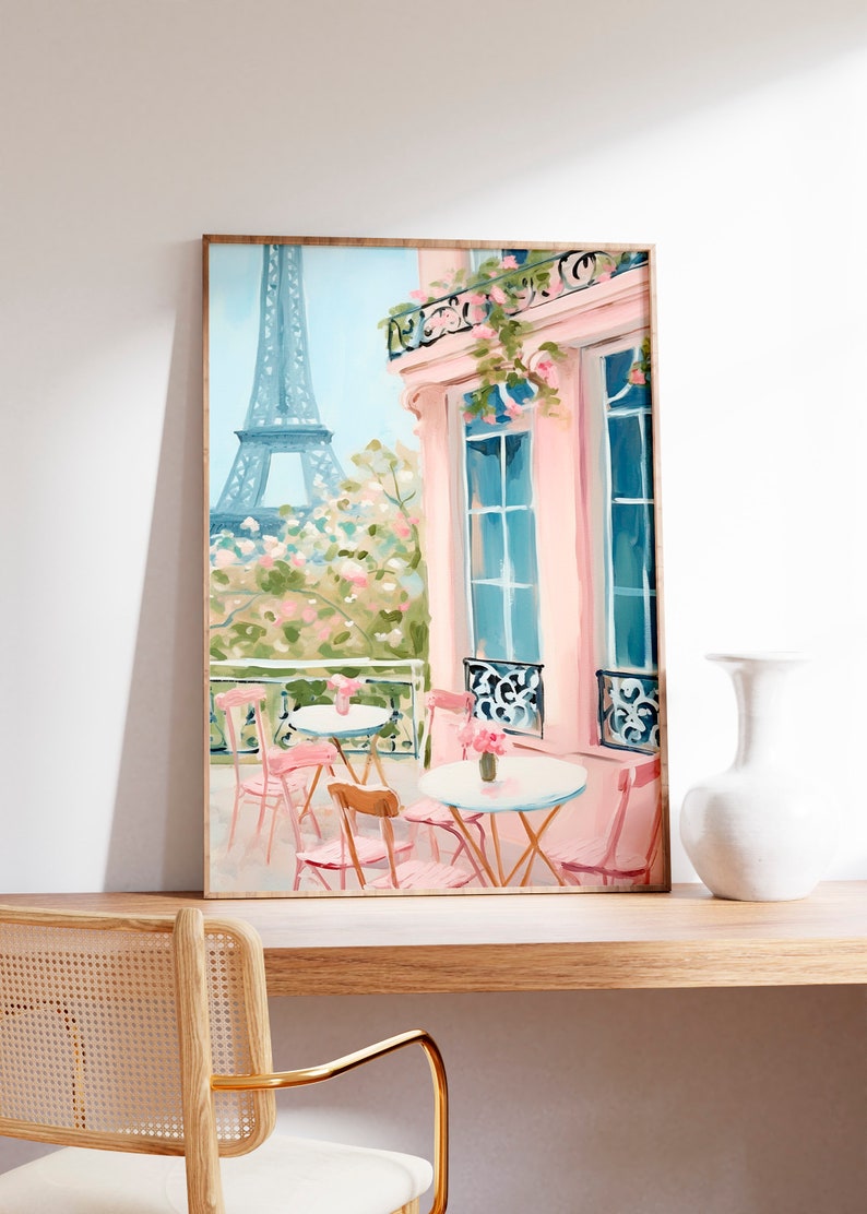 May include: A painting of a Parisian cafe with pink tables and chairs on a balcony overlooking the Eiffel Tower. The building is painted in a soft pink with blue windows and a balcony with pink flowers.