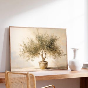 May include: A framed painting of an olive tree in a pot, with a light beige background. The painting is displayed on a wooden desk, next to a white vase. A wooden chair with a woven back is in the foreground.