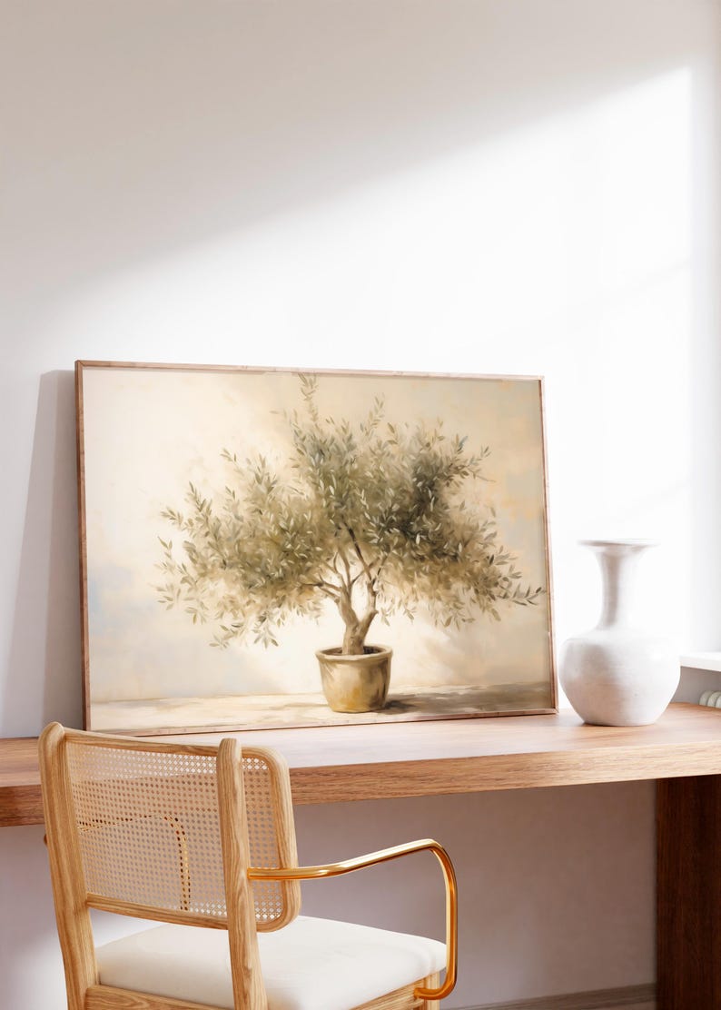 May include: A framed painting of an olive tree in a pot, with a light beige background. The painting is displayed on a wooden desk, next to a white vase. A wooden chair with a woven back is in the foreground.