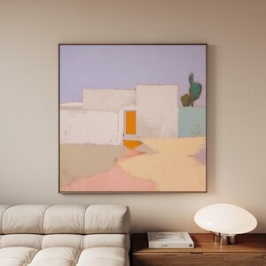 May include: Abstract painting featuring a minimalist architectural scene. White geometric shapes with an orange door are set against a lavender sky and a yellow and pink landscape. A green cactus adds a natural element. The artwork is framed.