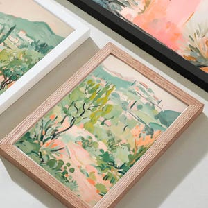 May include: Framed landscape art prints featuring a vibrant, impressionistic style. The artwork showcases a scene with green trees, a winding path, and buildings, with a color palette of greens, pinks, and oranges. The frames are in white, black, and natural wood.