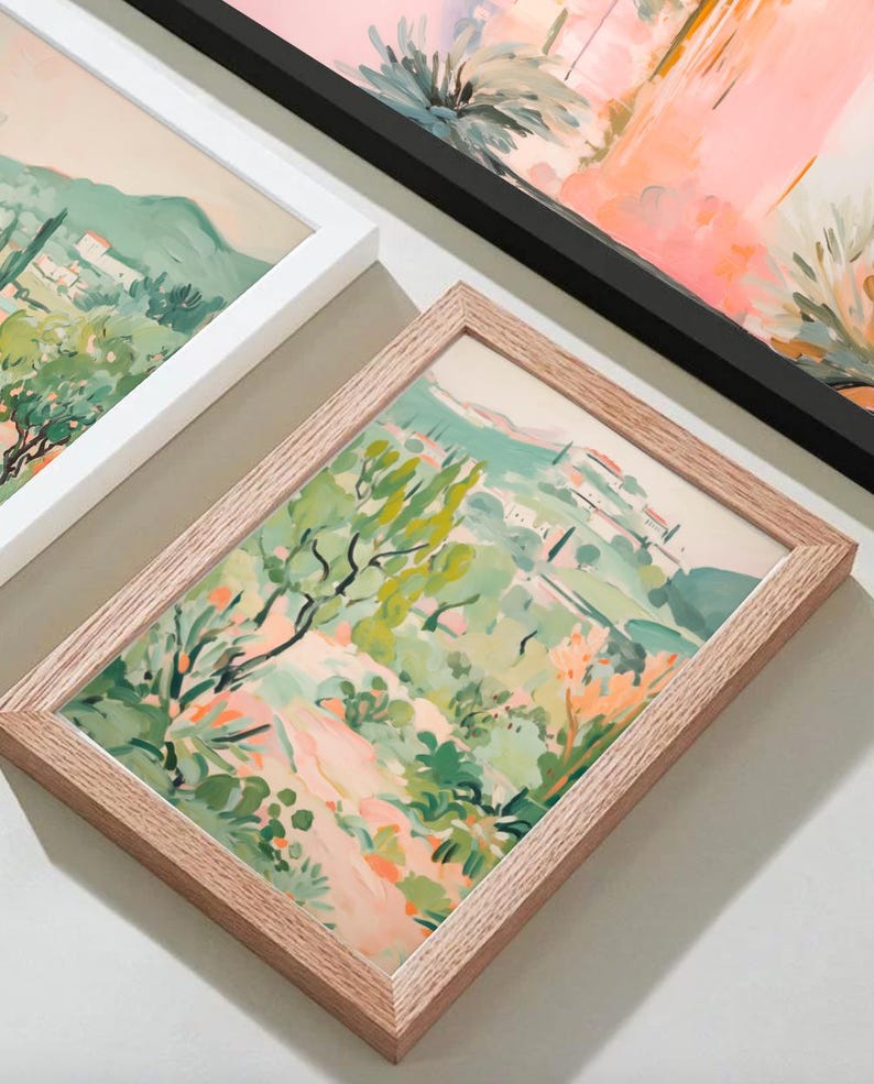 May include: Framed landscape art prints featuring a vibrant, impressionistic style. The artwork showcases a scene with green trees, a winding path, and buildings, with a color palette of greens, pinks, and oranges. The frames are in white, black, and natural wood.