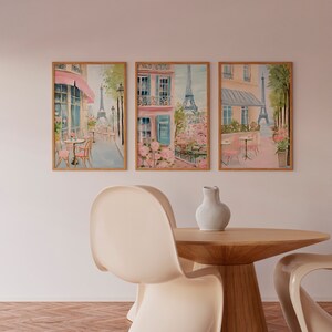 May include: Three framed watercolor paintings depicting Parisian street scenes with pink buildings, blooming trees, and the Eiffel Tower in the background. The paintings are hung on a white wall above a wooden table with two white chairs.