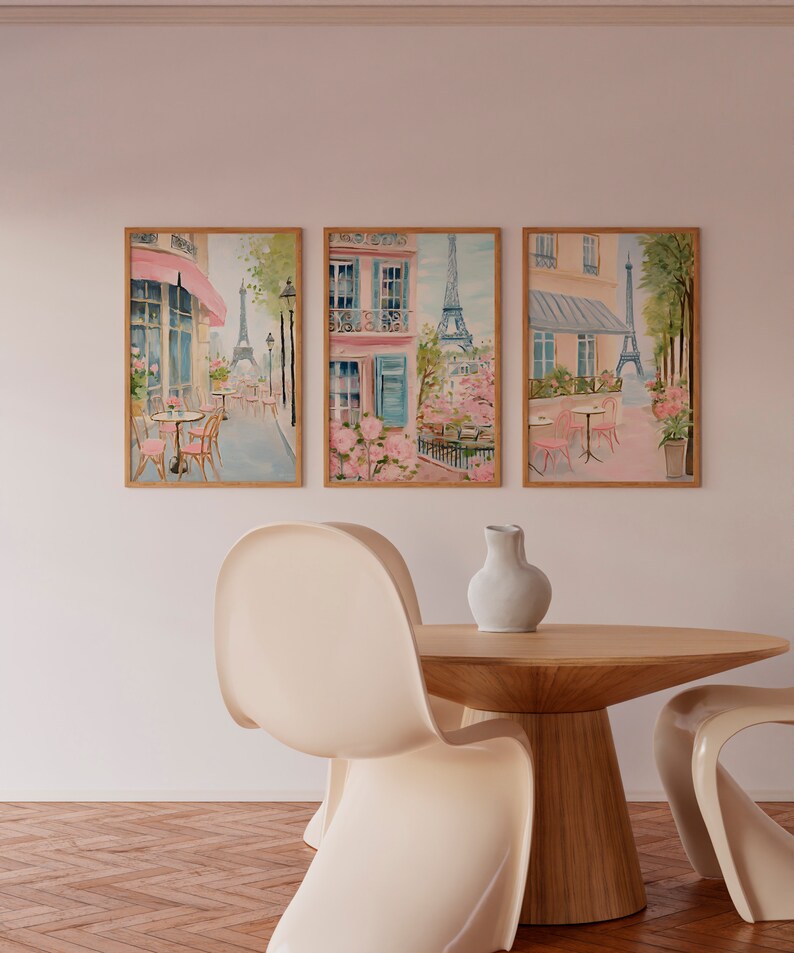 May include: Three framed watercolor paintings depicting Parisian street scenes with pink buildings, blooming trees, and the Eiffel Tower in the background. The paintings are hung on a white wall above a wooden table with two white chairs.