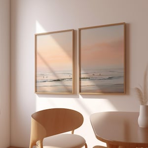 May include: Two framed beach landscape prints. The artwork depicts a serene ocean scene with surfers and a pastel-colored sky. The frames are a light wood color, and the overall aesthetic is calming and coastal.