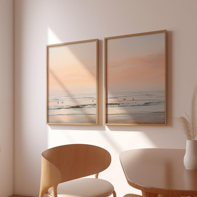 May include: Two framed beach landscape prints. The artwork depicts a serene ocean scene with surfers and a pastel-colored sky. The frames are a light wood color, and the overall aesthetic is calming and coastal.