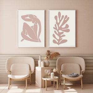 May include: Two framed art prints. One features a nude figure in a seated pose, the other a coral-like shape, both in a muted rose color against a cream background. The frames are white. The art is displayed in a room with matching furniture.