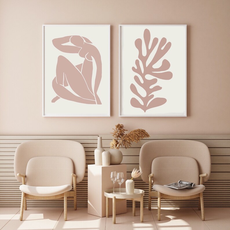 May include: Two framed art prints. One features a nude figure in a seated pose, the other a coral-like shape, both in a muted rose color against a cream background. The frames are white. The art is displayed in a room with matching furniture.