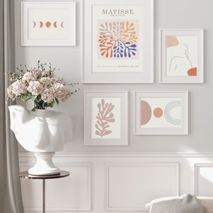 May include: A collection of framed art prints in white frames. The prints feature abstract designs, including a Matisse-inspired piece with colorful shapes, a line drawing of a figure, and a series of moon phases.