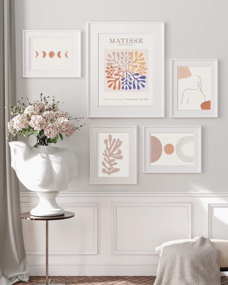 May include: A collection of framed art prints in white frames. The prints feature abstract designs, including a Matisse-inspired piece with colorful shapes, a line drawing of a figure, and a series of moon phases.
