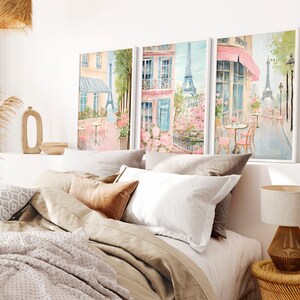 May include: Three framed watercolor paintings depicting Parisian street scenes with pink and blue hues. The paintings feature cafes, flowers, and the Eiffel Tower.