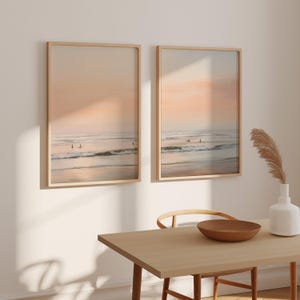 May include: Two framed beach art prints with a serene ocean scene. The artwork features a soft color palette of peach, beige, and blue, with surfers in the distance. The frames are light wood, and the prints are displayed on a white wall.