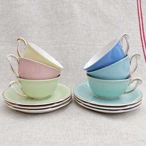 Könnte beinhalten: Sechs pastellfarbene Teetassen und Untertassen. Die Tassen sind paarweise gestapelt, wobei jedes Paar eine andere Farbe aufweist: Gelb, Rosa, Grün, Hellblau und Türkis. Die Untertassen haben einen gewellten Rand und einen goldenen Rand. Die Griffe sind weiß mit goldenen Akzenten.