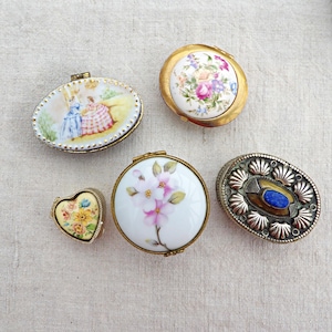 May include: Five small trinket boxes including an oval box with a painted scene of two figures, a round box with floral patterns, a heart-shaped box with yellow flowers, a round box with pink blossoms, and an oval box with a blue stone inlay.