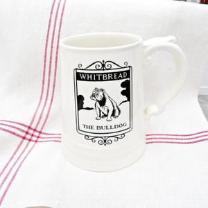 May include: A white ceramic mug with a curved handle. The mug features a black and white graphic of a bulldog and the text "WHITBREAD THE BULLDOG" within a decorative frame. The mug is on a white and red striped cloth.