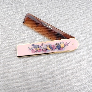 Peut inclure: Un peigne de poche pliable avec un peigne en plastique brun imitation écaille de tortue et un étui en métal rose clair. L'étui présente une illustration décorative de quatre petits oiseaux sur une branche fleurie et la marque Stratton en argent.