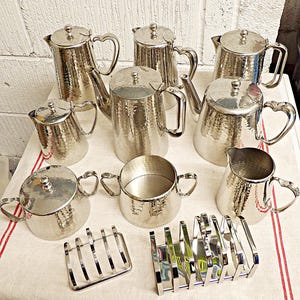 May include: A collection of silver-toned hammered metal tea service pieces including three tall pots, two smaller pitchers, a lidded sugar bowl, an open sugar bowl, a creamer, and two metal toast racks, all featuring heart-shaped handle accents.