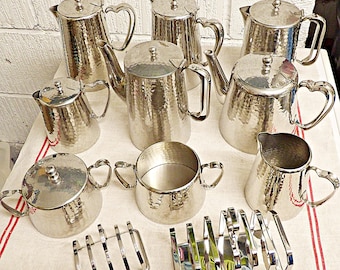 May include: A collection of silver-toned hammered metal tea service pieces including three tall pots, two smaller pitchers, a lidded sugar bowl, an open sugar bowl, a creamer, and two metal toast racks, all featuring heart-shaped handle accents.