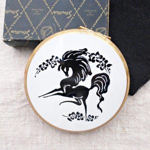 May include: A vintage Stratton compact mirror with a gold-tone rim and a white face. The face features a black silhouette of a horse with floral accents. The original box is visible in the background.