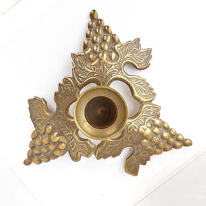 May include: A vintage brass candle holder with a grape and leaf design. The candle holder has a circular base and is surrounded by detailed grape clusters and leaf patterns. The brass has a warm, aged patina, and the overall shape is triangular.