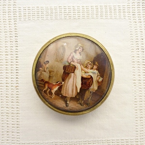 Op de afbeelding: Een ronde, goudkleurige knoop met een verhoogde glazen koepel. De koepel toont een geschilderd tafereel van een vrouw in een witte jurk en een mand, met twee kinderen en een hond op de achtergrond.