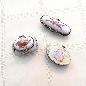 Könnte beinhalten: Drei kleine Porzellan-Schmuckkästchen mit goldenen Verzierungen und bemalten Blumen- und Fruchtmotiven. Die Kästchen sind oval und rechteckig.