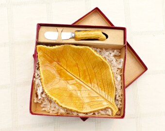 May include: A red box containing a yellow ceramic leaf-shaped dish and a small silver butter knife. The dish is shaped like a leaf with a raised vein pattern.