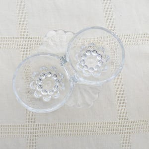 Op de afbeelding: Een dubbele glazen schaal met een gegolfde rand en een getextureerd midden. De schaal is ontworpen om sauzen of snacks te serveren.