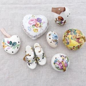 May include: A collection of white ceramic decorative items, including heart-shaped and oval-shaped pieces, and a pair of clogs. The items are adorned with floral and pastoral designs, and some have gold accents.