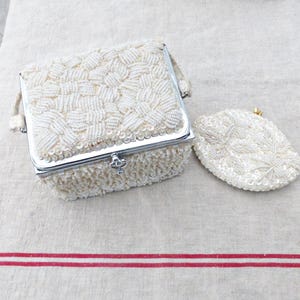 May include: A set of two white beaded purses. The larger purse has a hinged lid and a silver clasp. The smaller purse is a coin purse.