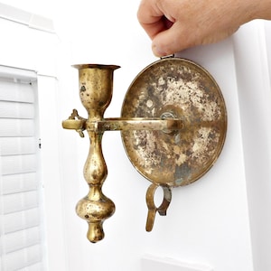 Op de afbeelding: Een vintage messing wandkaarsenhouder. De kaarsenhouder heeft een ronde achterplaat, een horizontale arm en een kopvormige houder voor de kaars. Het messing heeft een aangetaste, verouderde uitstraling, wat bijdraagt aan de antieke charme. Een decoratief stuk voor de interieurdecoratie.