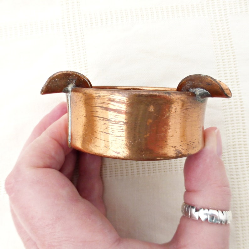 May include: A copper ashtray with two small, rounded, outward-facing wings on either side. The ashtray is slightly dented and has a worn, aged look.