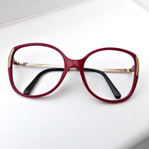 Op de afbeelding: Rode plastic gemonteerde bril met gouden accenten en zwarte pootjes.